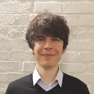 Smiling person with dark hair and sweater in front of a white brick wall